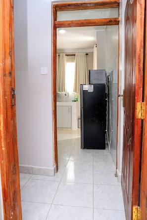 A hallway leading to a kitchen with a refrigerator and a window with curtains. - Cosy 1BR Apartment with Balcony, Free Wi-Fi & Secure Parking (Nairobi)