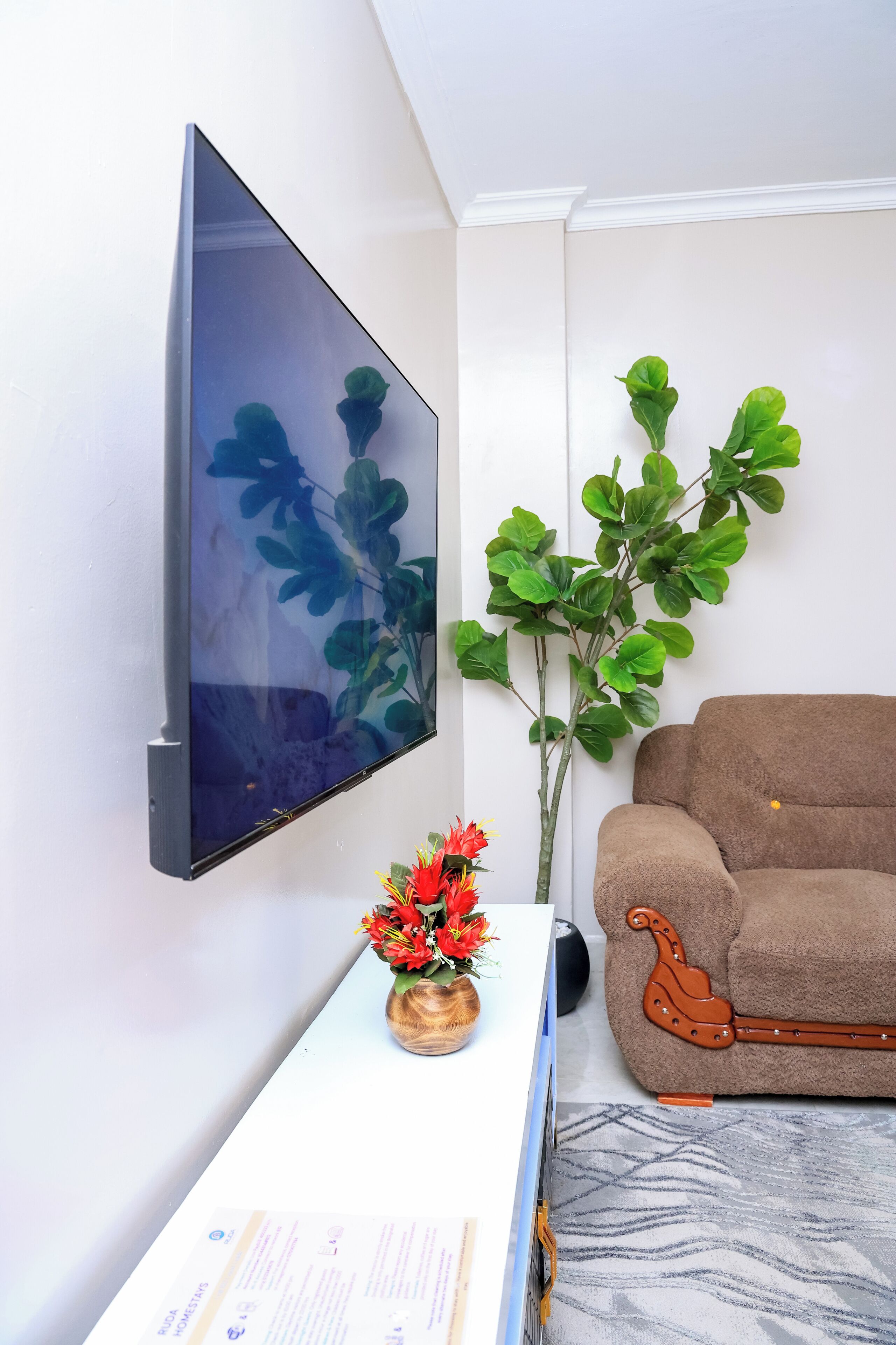 A modern living room with a flat-screen TV mounted on the wall, a potted plant, a brown sofa, and a wooden side table with a vase of flowers.