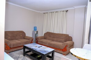 A living room with two brown sofas, a marble coffee table, and a water dispenser. - Cosy 1BR Apartment with Balcony, Free Wi-Fi & Secure Parking (Nairobi)