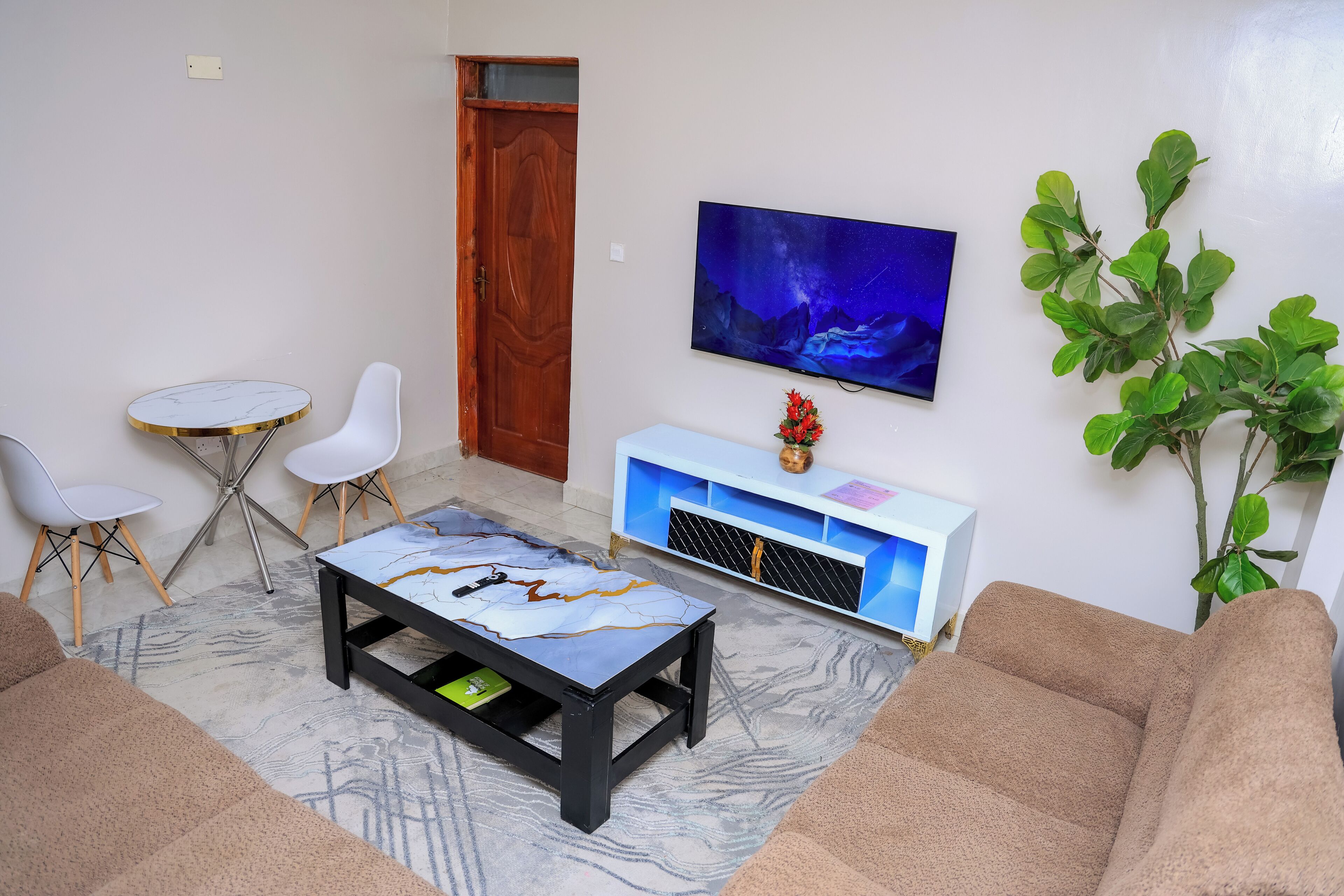 Una sala de estar con una mesa de centro de mármol, un televisor montado en la pared, un armario blanco y una planta verde grande.