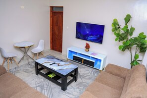 A living room with a marble coffee table, a television mounted on the wall, a white cabinet, and a large green plant. - Cosy 1BR Apartment with Balcony, Free Wi-Fi & Secure Parking (Nairobi)