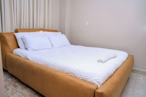 A bed with a brown headboard and white bedding in a room with a plain wall and a light-colored floor. - Cosy 1BR Apartment with Balcony, Free Wi-Fi & Secure Parking (Nairobi)