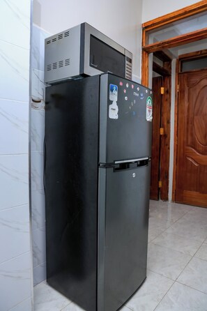 A black refrigerator with a microwave on top, a door with stickers, and a tiled floor. - Cosy 1BR Apartment with Balcony, Free Wi-Fi & Secure Parking (Nairobi)