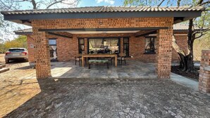 Un patio techado al aire libre con pared de ladrillo, una mesa con sillas y un coche estacionado al fondo.