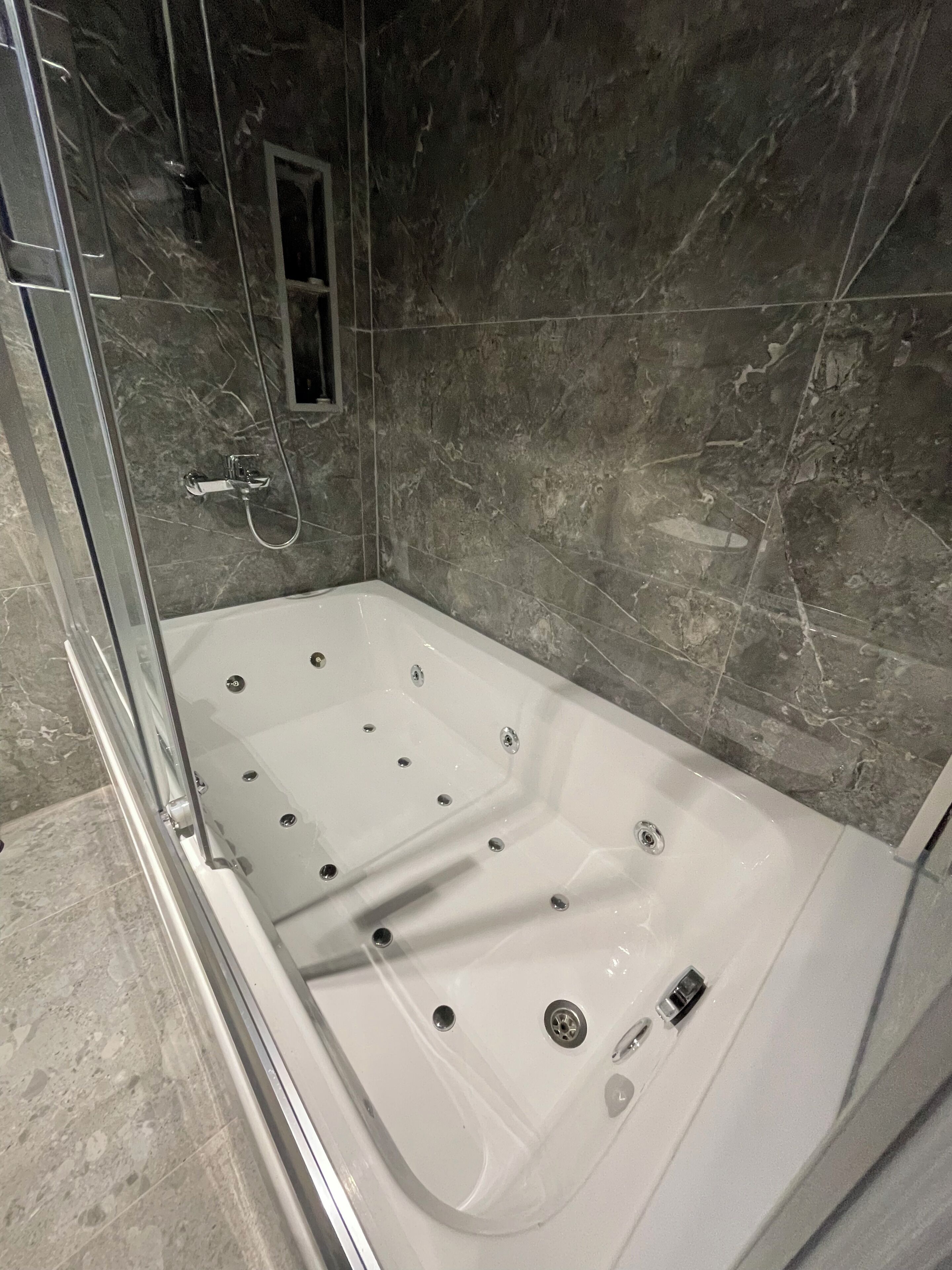 Un baño moderno con ducha envuelta en vidrio, una bañera blanca con varios grifos y una pared de mármol.