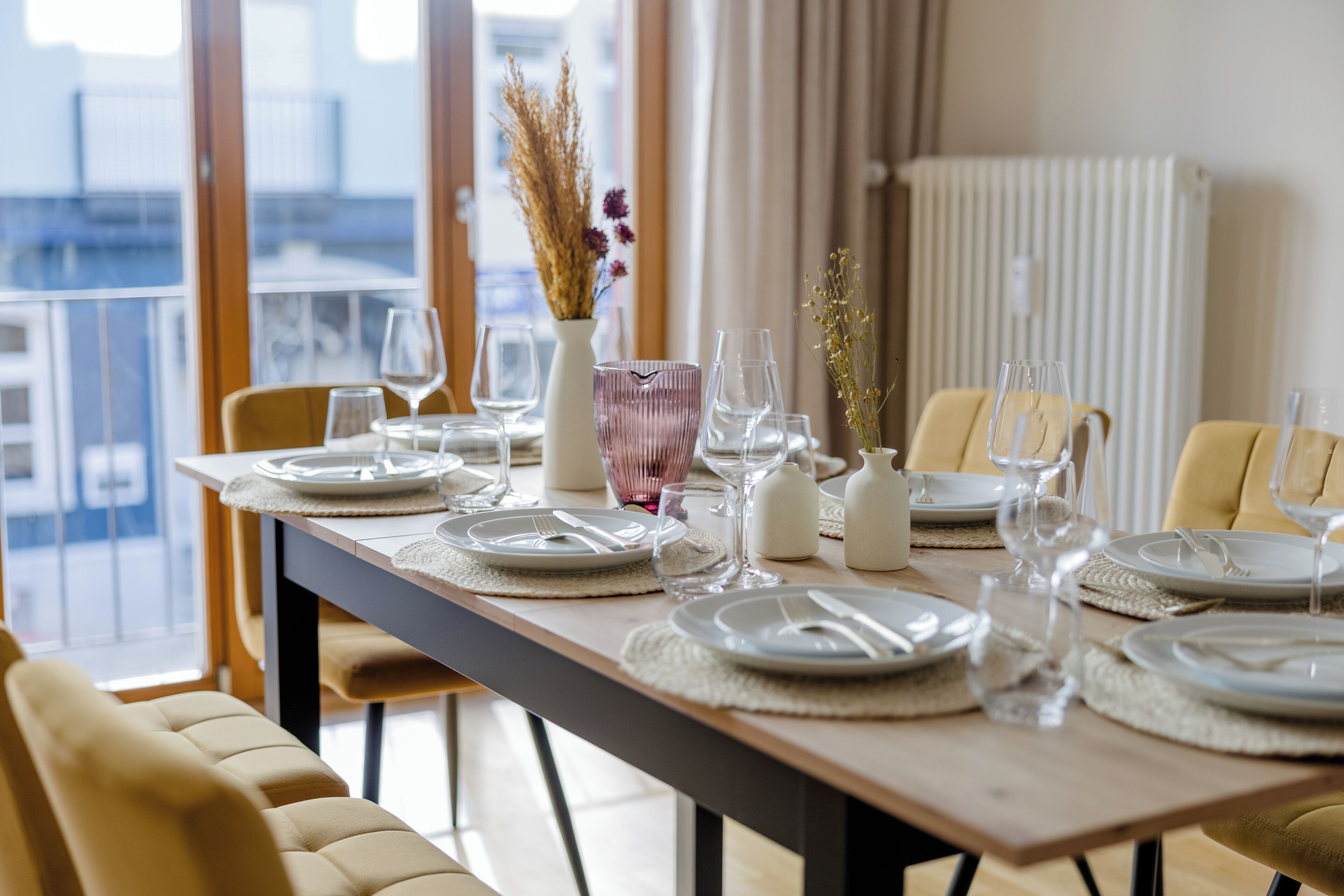 Una mesa de comedor dispuesta con platos, cubiertos y vasos, con vistas a la ciudad a través de grandes ventanales.