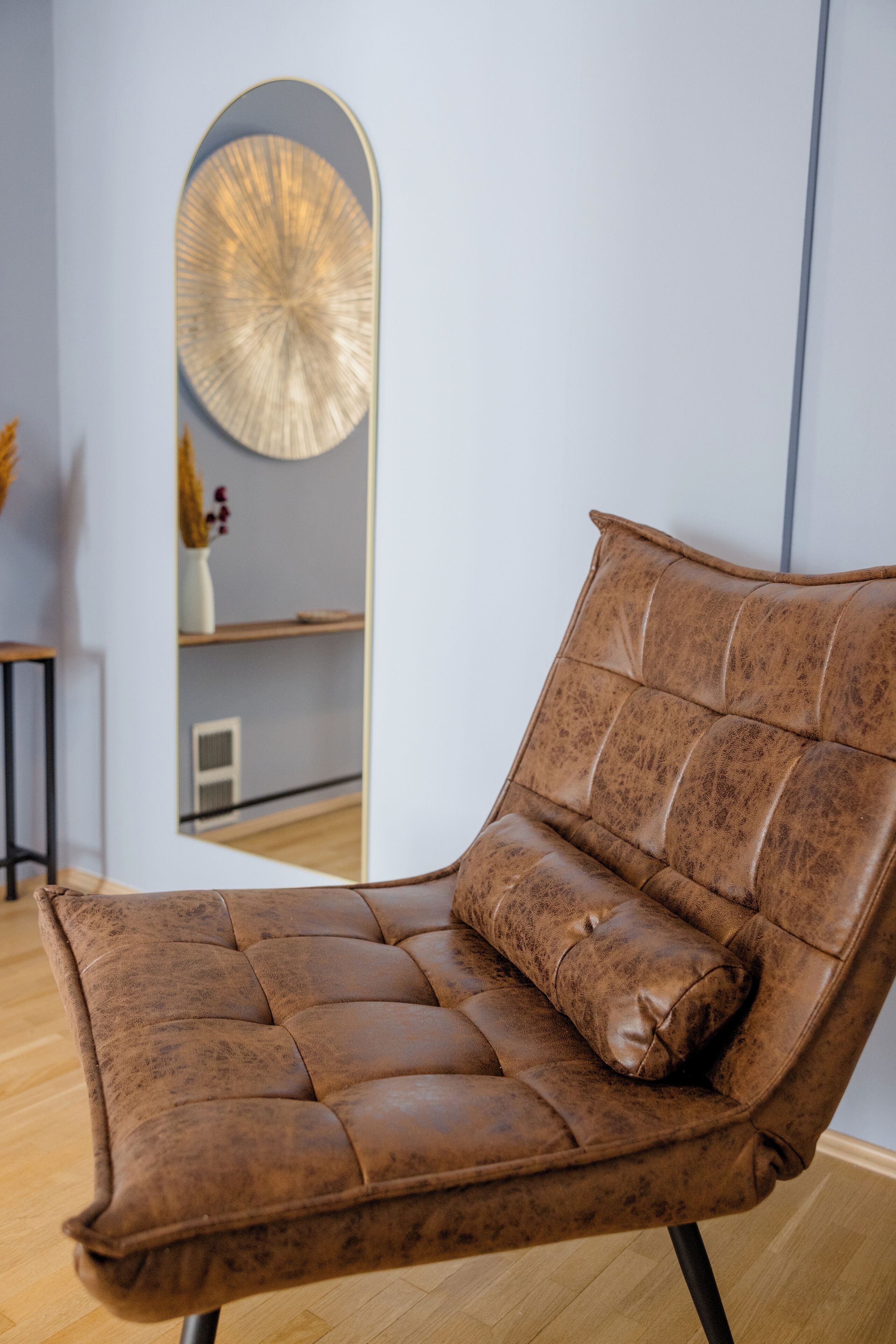 Un fauteuil en cuir marron avec un coussin, placé dans une pièce dont le sol est en bois et où l’on trouve un grand miroir au mur.