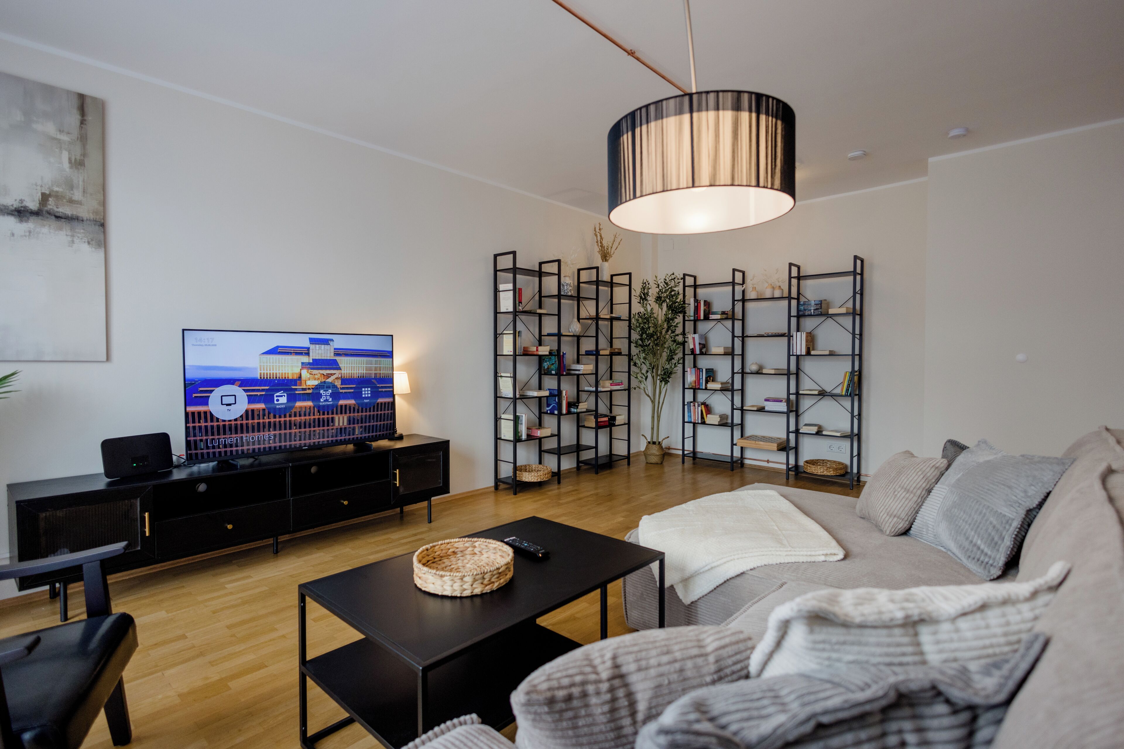 A modern living room with a flat-screen TV, a black entertainment unit, a large black coffee table, a grey sofa, and a decorative plant.