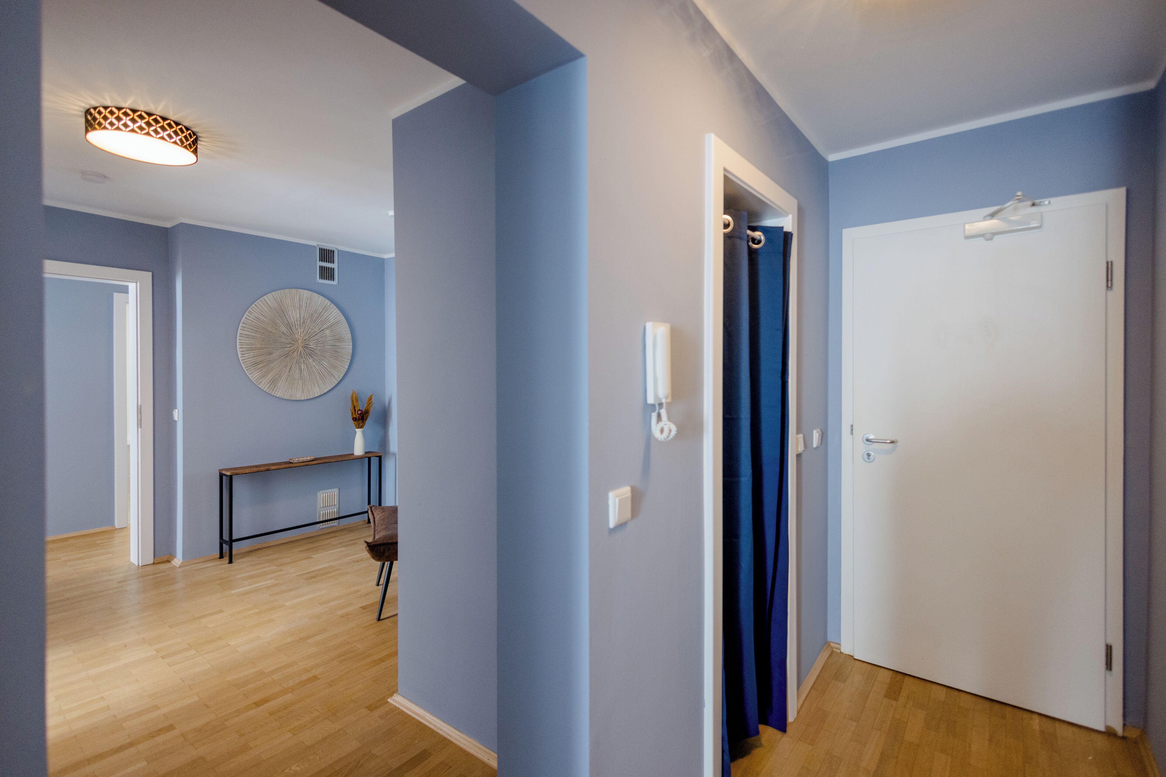 Un pasillo con piso de madera, un adorno circular en la pared, una mesita con un florero y una puerta con cortina.