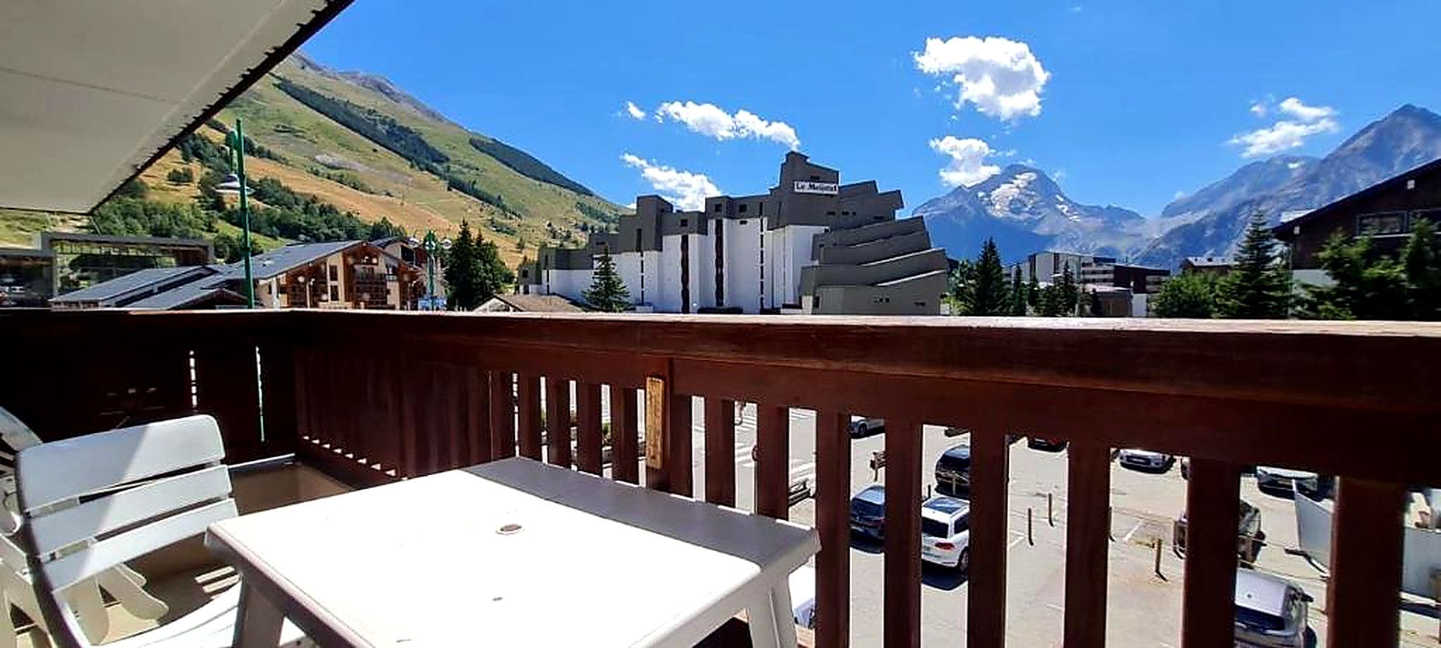 A balcony with a table and chairs, overlooking a street with parked cars and a mountain in the background.