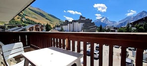 Un balcon avec une table et des chaises, donnant sur une rue bordée de voitures garées et surmontée d’une montagne en arrière-plan.