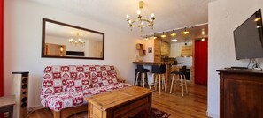 A living room with a sofa, a wooden coffee table, a TV, and a kitchen area in the background. - Charming apartment for 6 people with a mountain view (Les Deux Alpes)