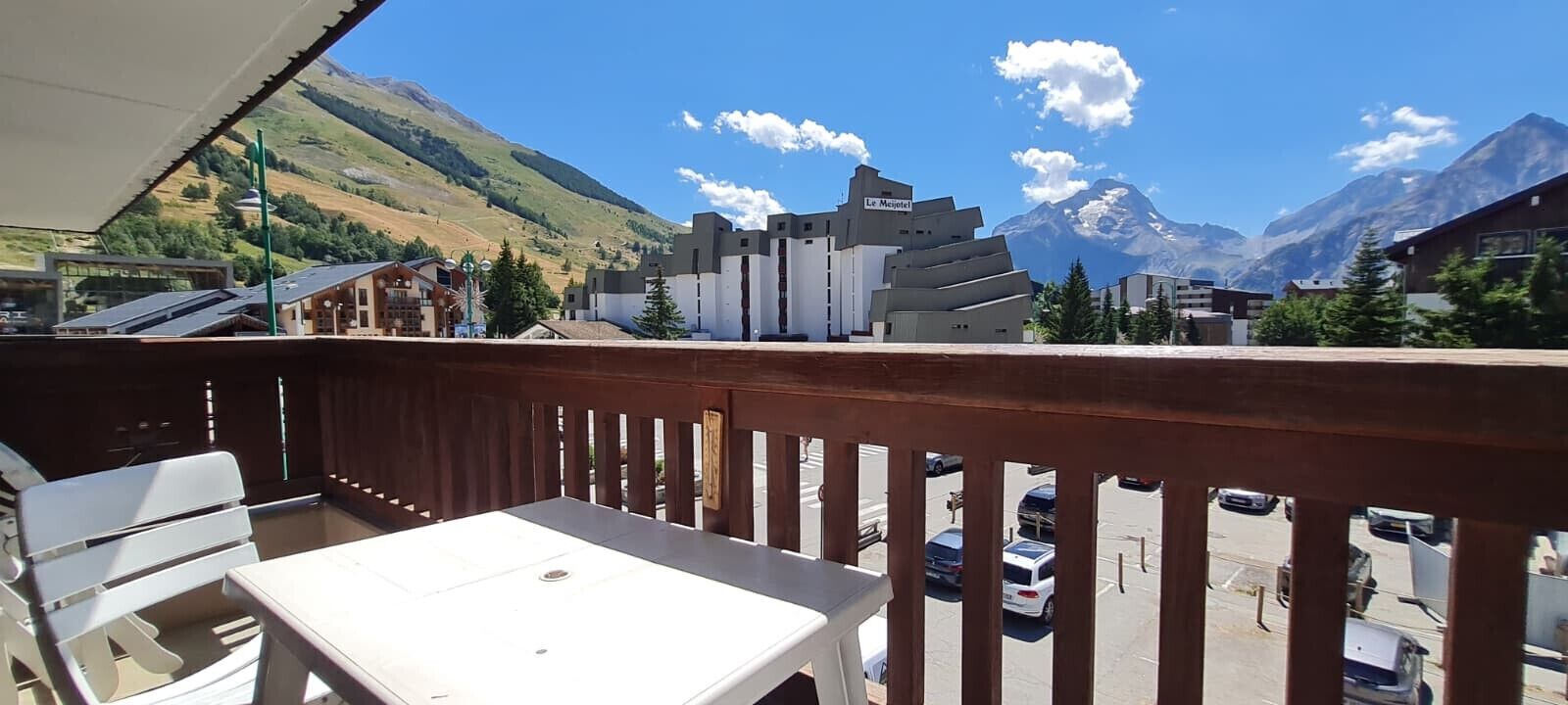 A balcony with a table and chairs, overlooking a parking area and buildings with a mountain backdrop.