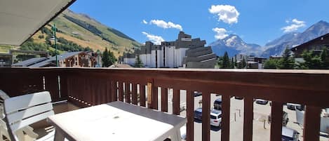 A balcony with a table and chairs, overlooking a parking area and buildings with a mountain backdrop.