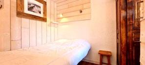 Uma cama de solteiro com colchão branco e cabeceira de madeira, um banquinho de madeira pequeno e um quadro emoldurado na parede.