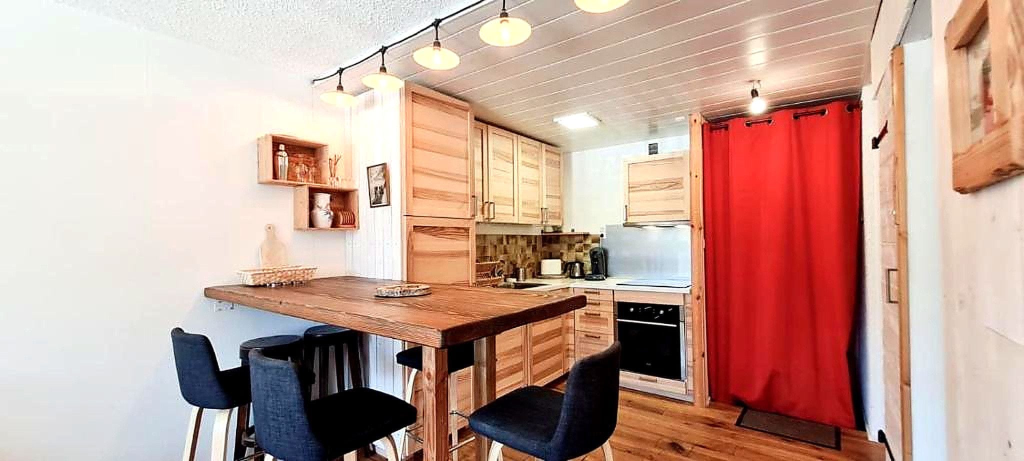 A compact kitchen with wooden cabinets, a dining table with four chairs, and a red curtain.