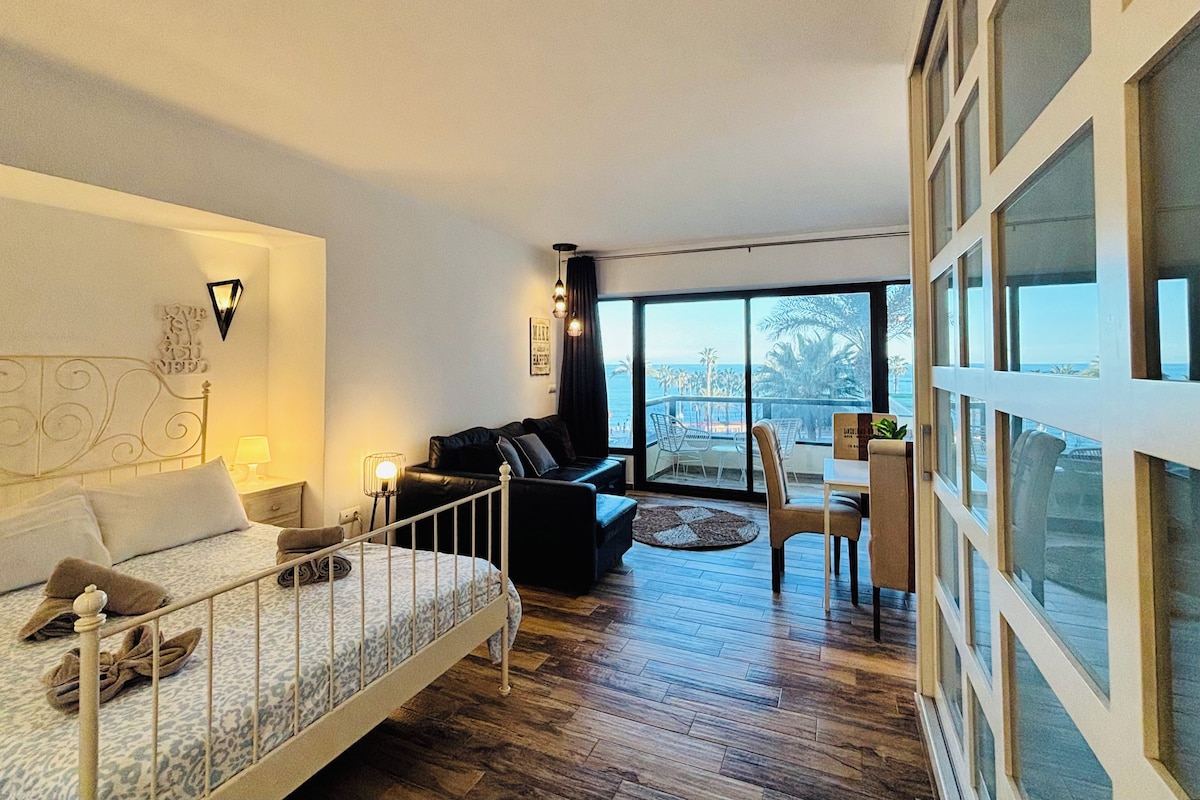 A hotel room with a bed, a sofa, a dining area, and a view of the sea.