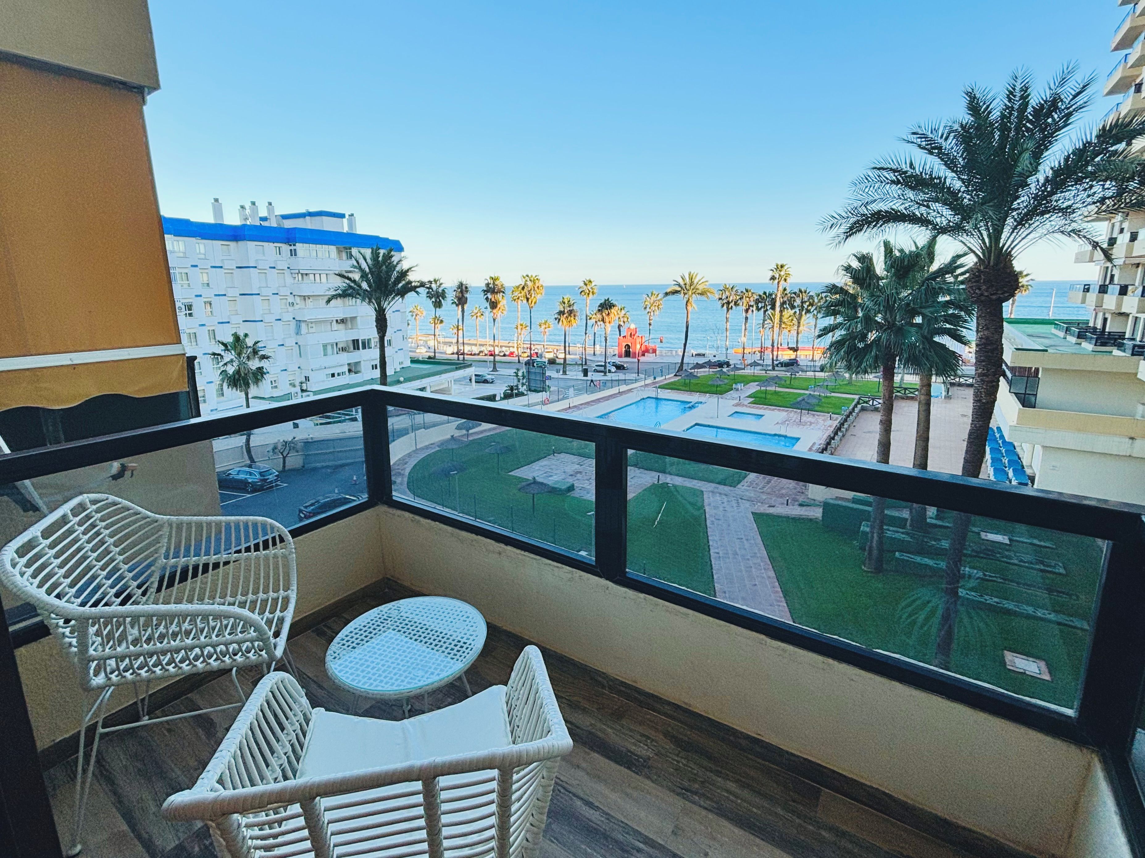 A balcony with wicker furniture, a pool, and a view of palm trees and buildings.