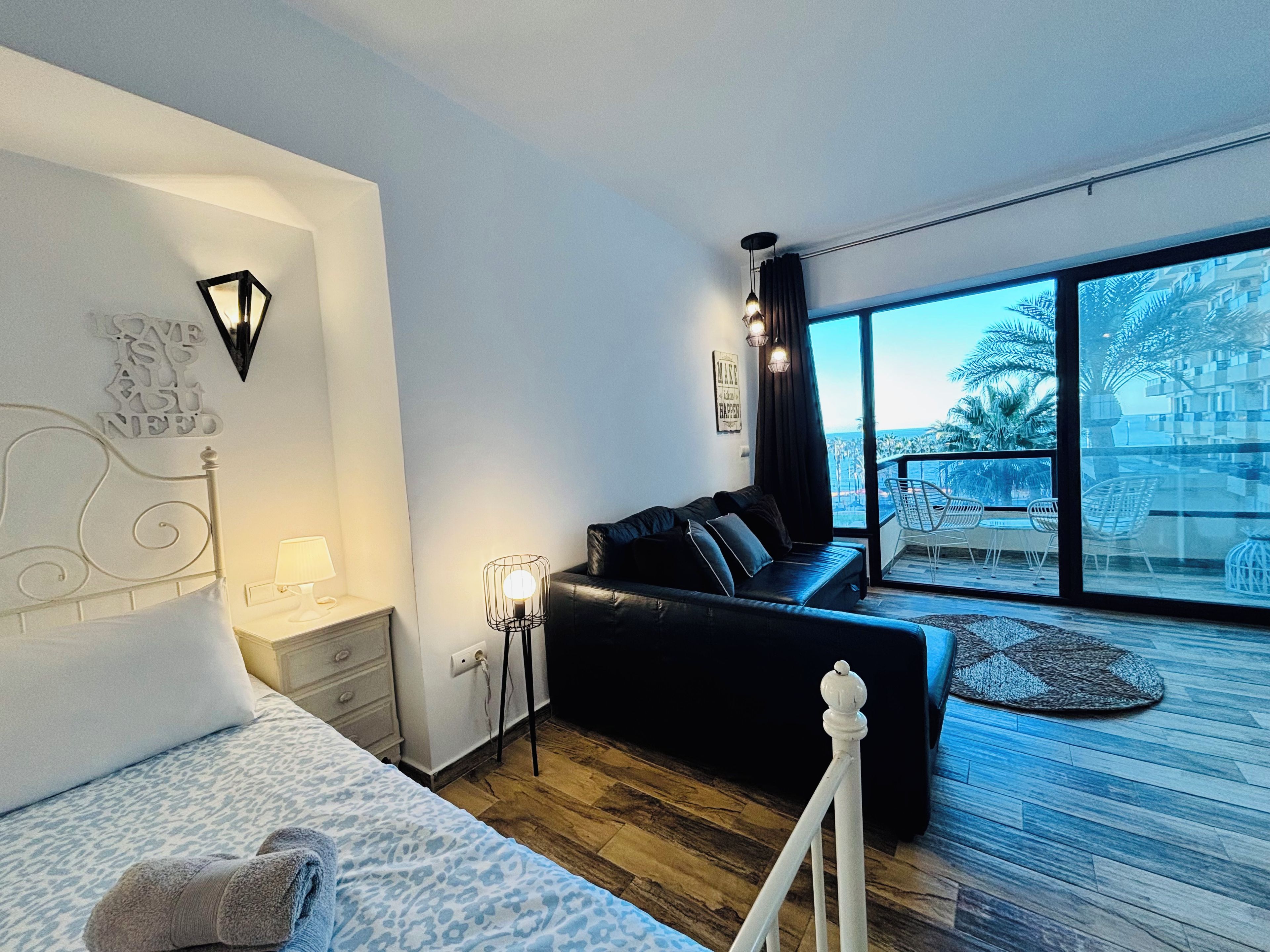Une chambre moderne avec un lit, un canapé, une table de chevet, une lampe et une grande fenêtre donnant sur des palmiers et des immeubles.
