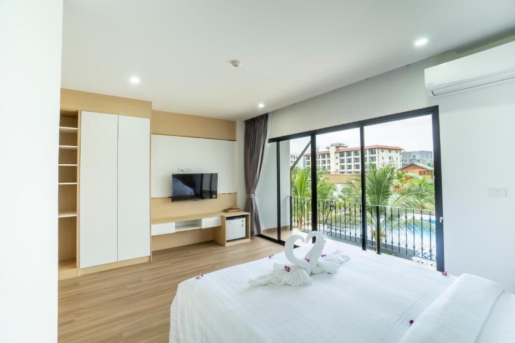 A hotel room with a bed, a TV, a wardrobe, and a balcony with a view of buildings and greenery.