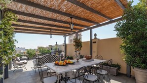A covered outdoor patio with a table and chairs, surrounded by potted plants and a view of the cityscape. - Casa Dolce Roma – Your home in the heart of Rome! (Roma)