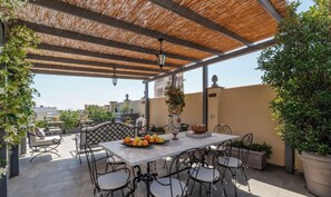 Eine überdachte Terrasse im Freien mit Tisch und Stühlen, umgeben von Blumentöpfen und Blick auf die Stadtlandschaft.