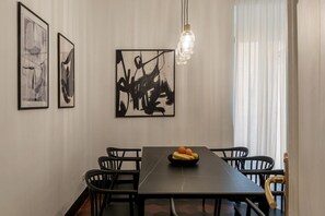 Ein Essbereich mit einem langen Tisch, schwarzen Stühlen und einer Obstschale. An der Wand hängen abstrakte Gemälde und über dem Tisch hängt eine Pendelleuchte.