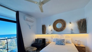Un dormitorio con cama, mesitas de noche, una silla, ventilador de techo y vista al océano.