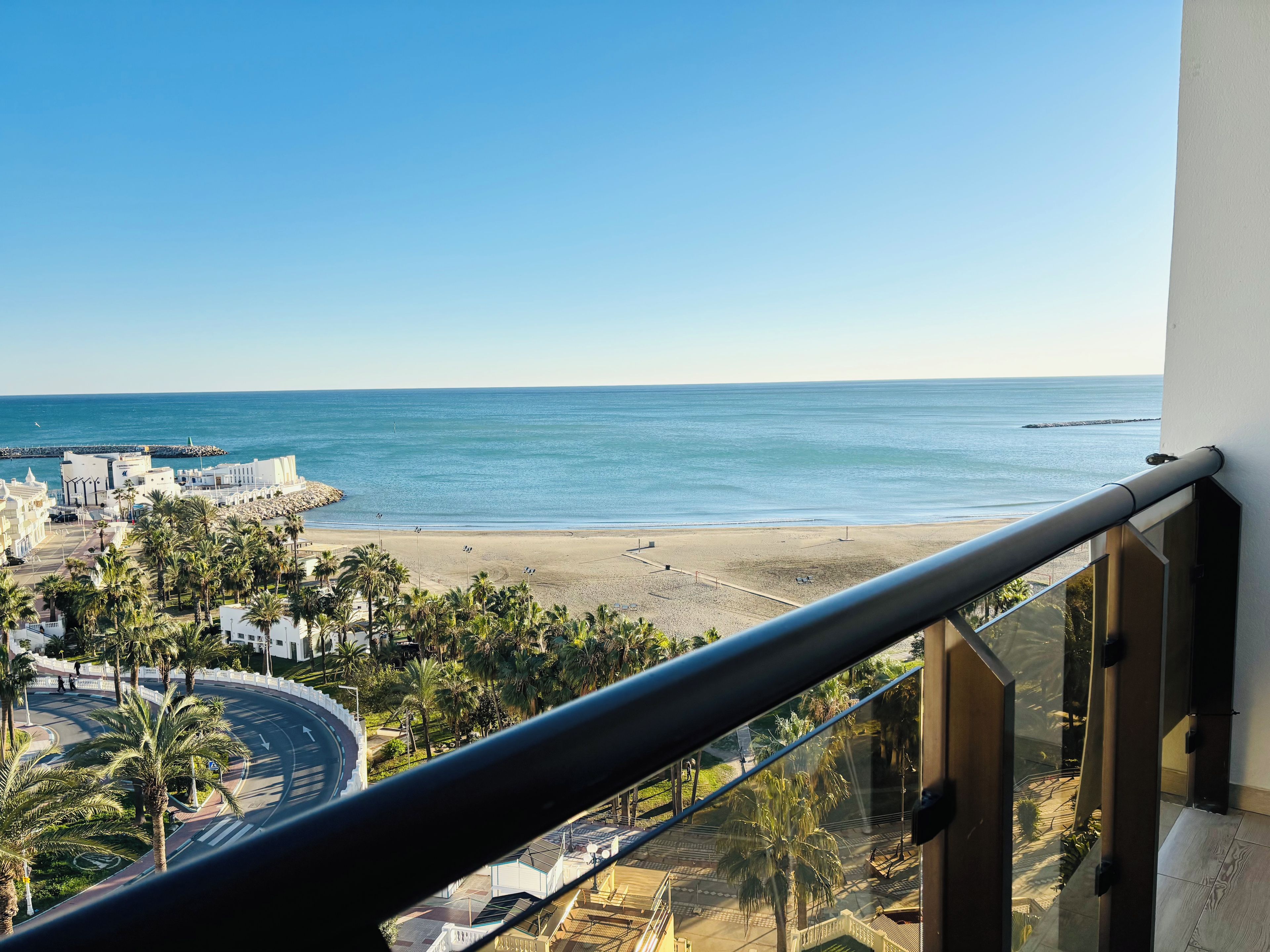A balcony with a view of a beach, palm trees, and a road with a roundabout.