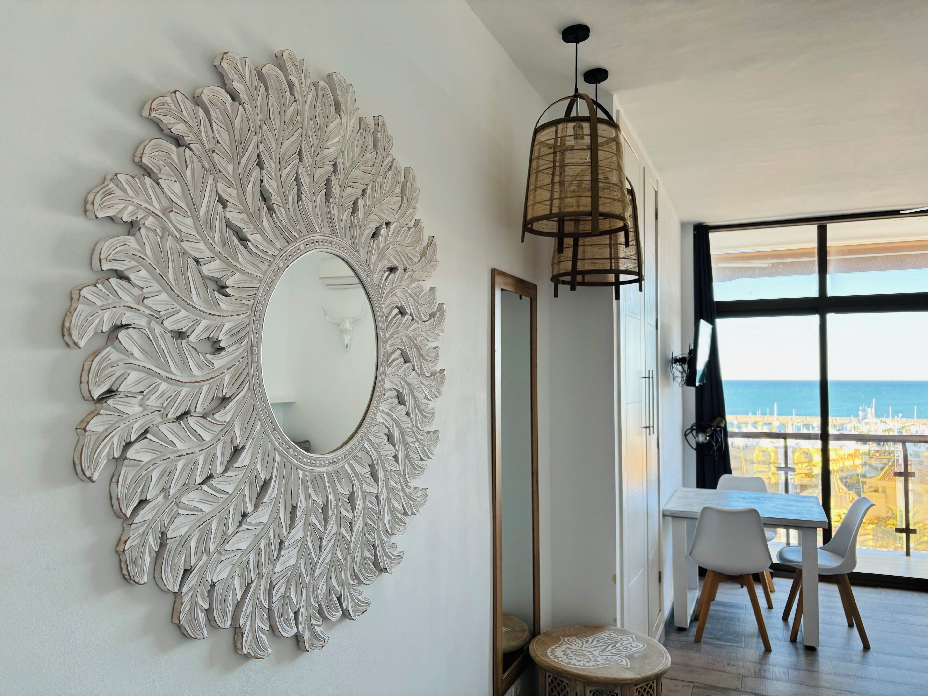 A room with a large decorative mirror, a dining table with chairs, and a view of the sea through large windows.