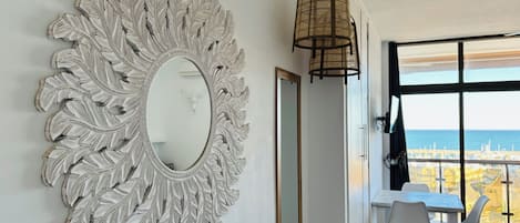 A room with a large decorative mirror, a dining table with chairs, and a view of the sea through large windows.