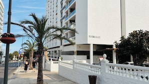 Un edificio moderno con fachada blanca y balcones, un paseo peatonal y palmeras.