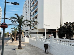 Um edifício moderno com fachada branca e varandas, uma passarela para pedestres e palmeiras.