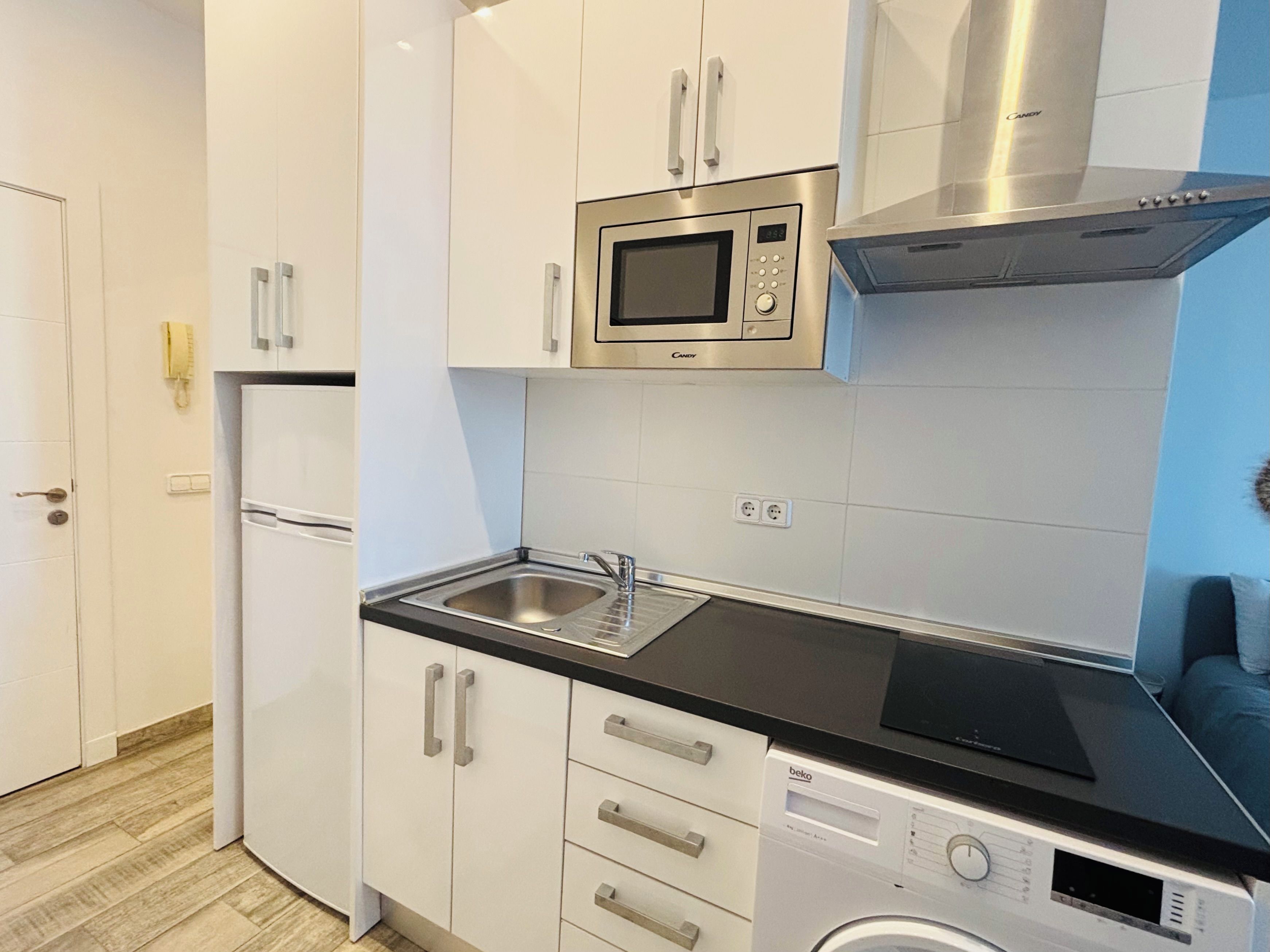 A modern kitchen with white cabinets, a stainless steel microwave, a built-in oven, a sink, and a washing machine.