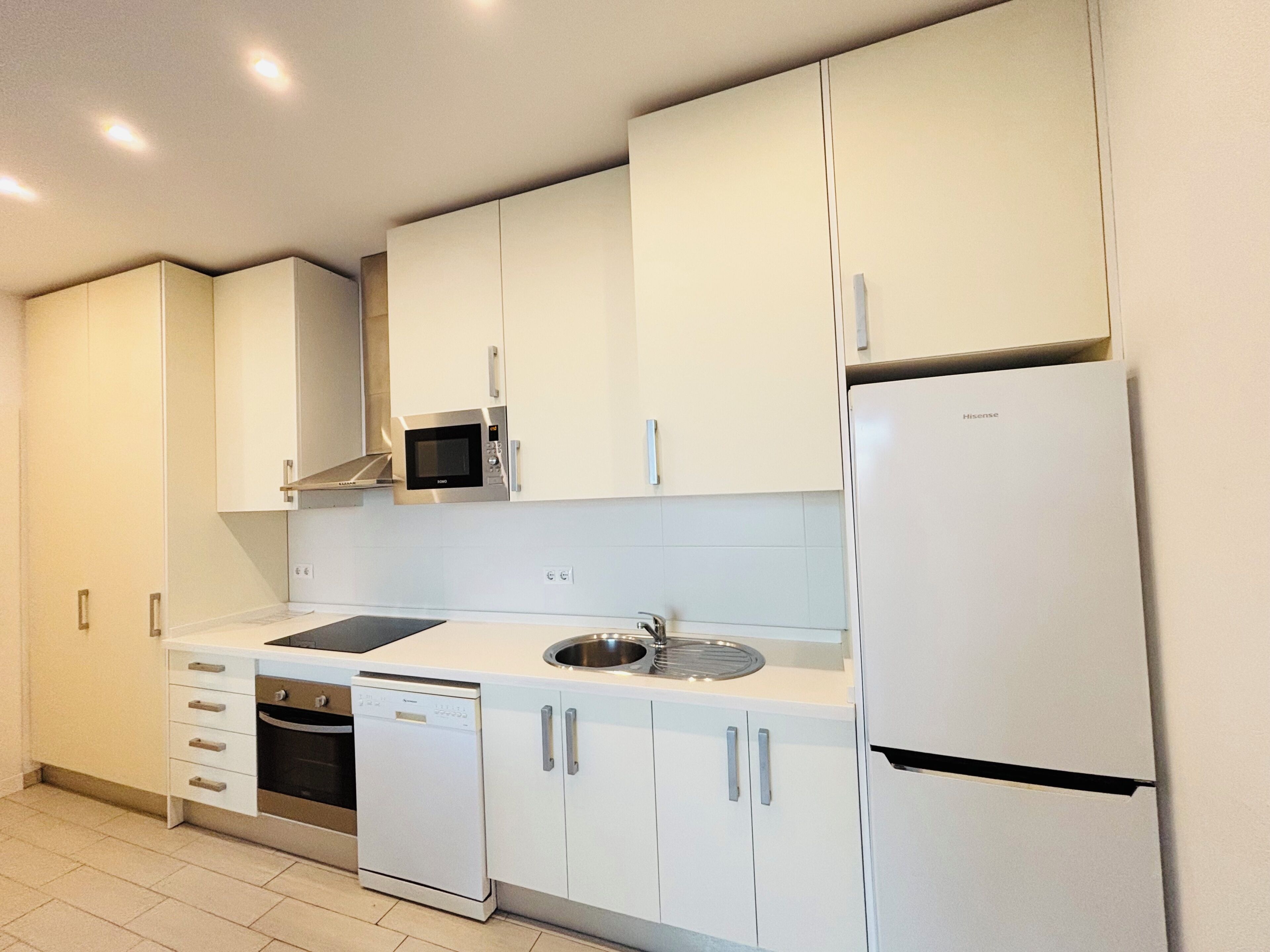 Una cocina moderna con armarios blancos, microondas integrado, lavavajillas y refrigerador.