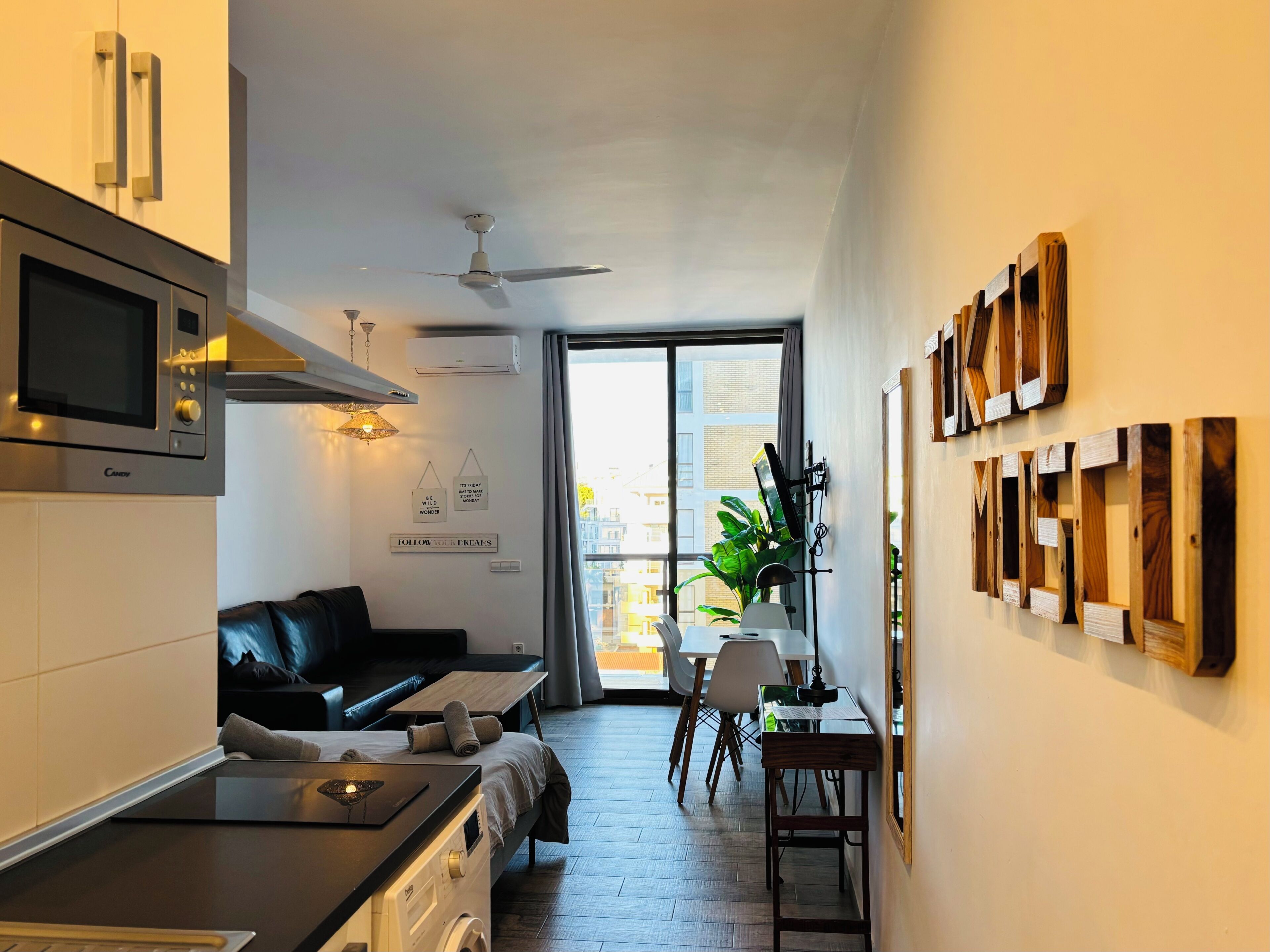 A modern kitchen with a microwave, stove, and ceiling fan. There is a dining area with a table and chairs, and a living space with a sofa.