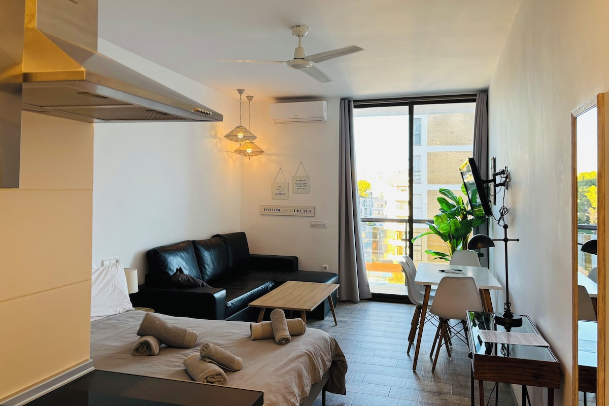 Quarto de hotel moderno com cama, sofá de couro preto, mesa de jantar e uma janela grande com vista para edifícios.