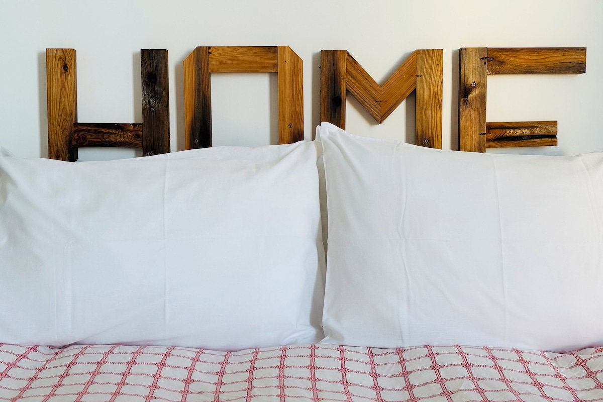 Uma cama com lençóis brancos e um cobertor estampado, um travesseiro, e as letras de madeira 'HOME' na parede.