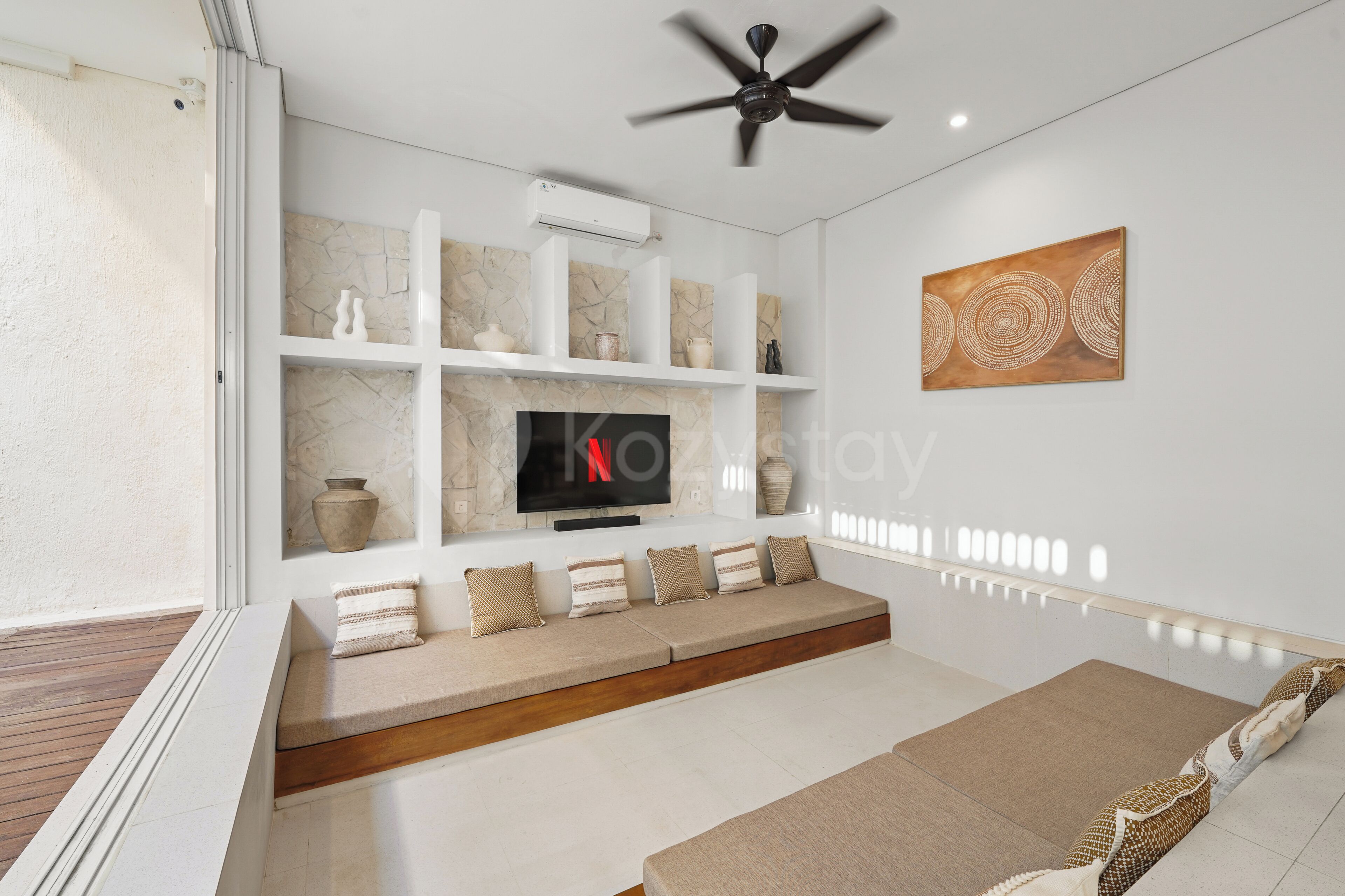 A modern living room with a flat-screen TV, built-in shelves, a ceiling fan, and a decorative wall piece.