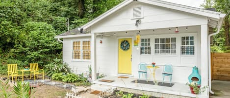 A white house with a yellow door, a porch with chairs, and a small garden.