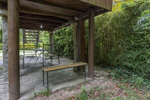 Una estructura de fusta a l'aire lliure amb bancs i un paviment de pedra, envoltada de densa vegetació de bambú.