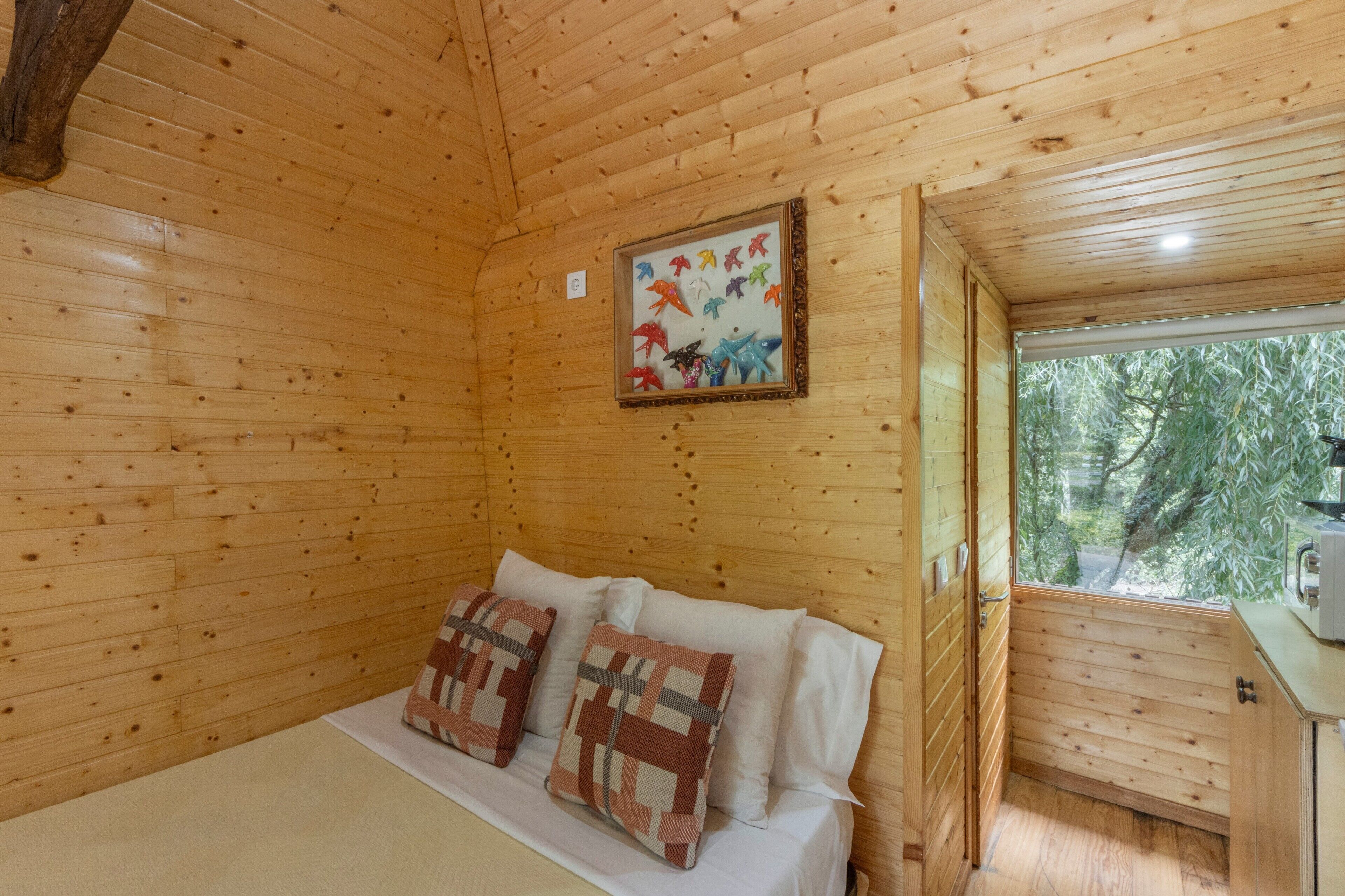 Ein Zimmer in einer Holzhütte mit einem Bett, Kissen, einem gerahmten Bild an der Wand und einem Fenster mit Blick auf Bäume.