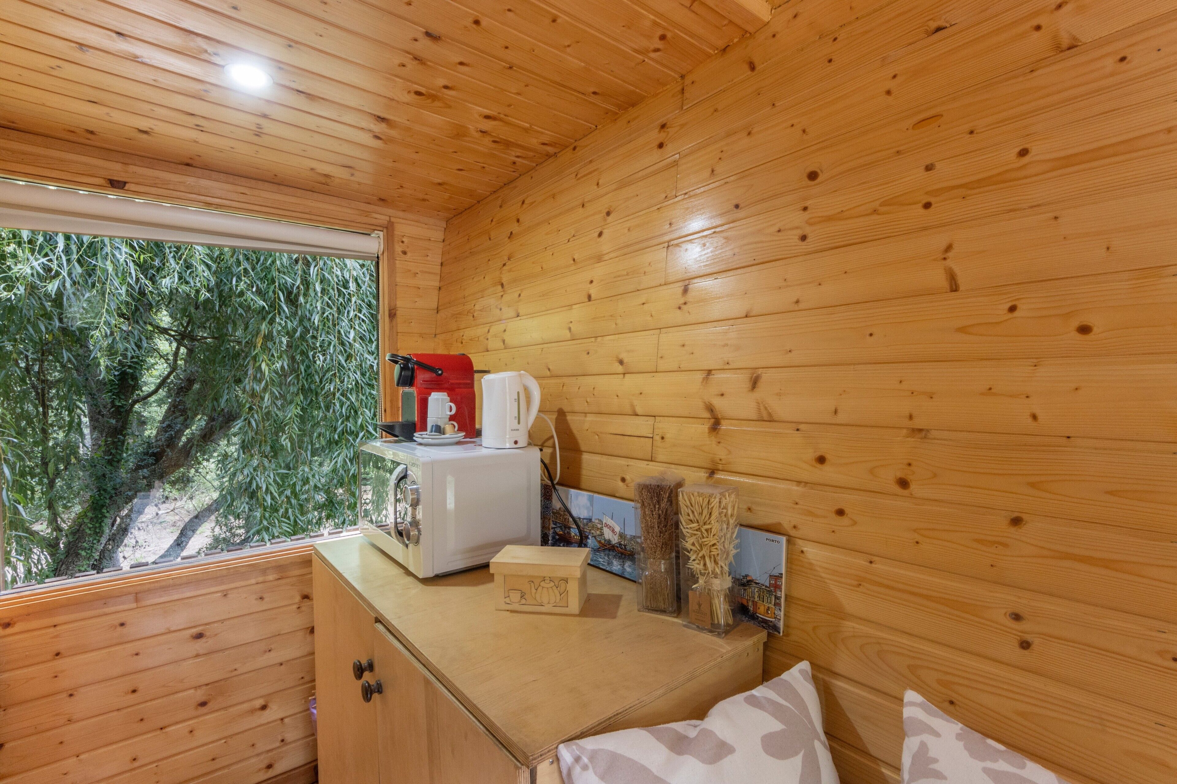 A wooden cabin interior with a kitchenette, a window overlooking greenery, and a small table with a cup and saucer.