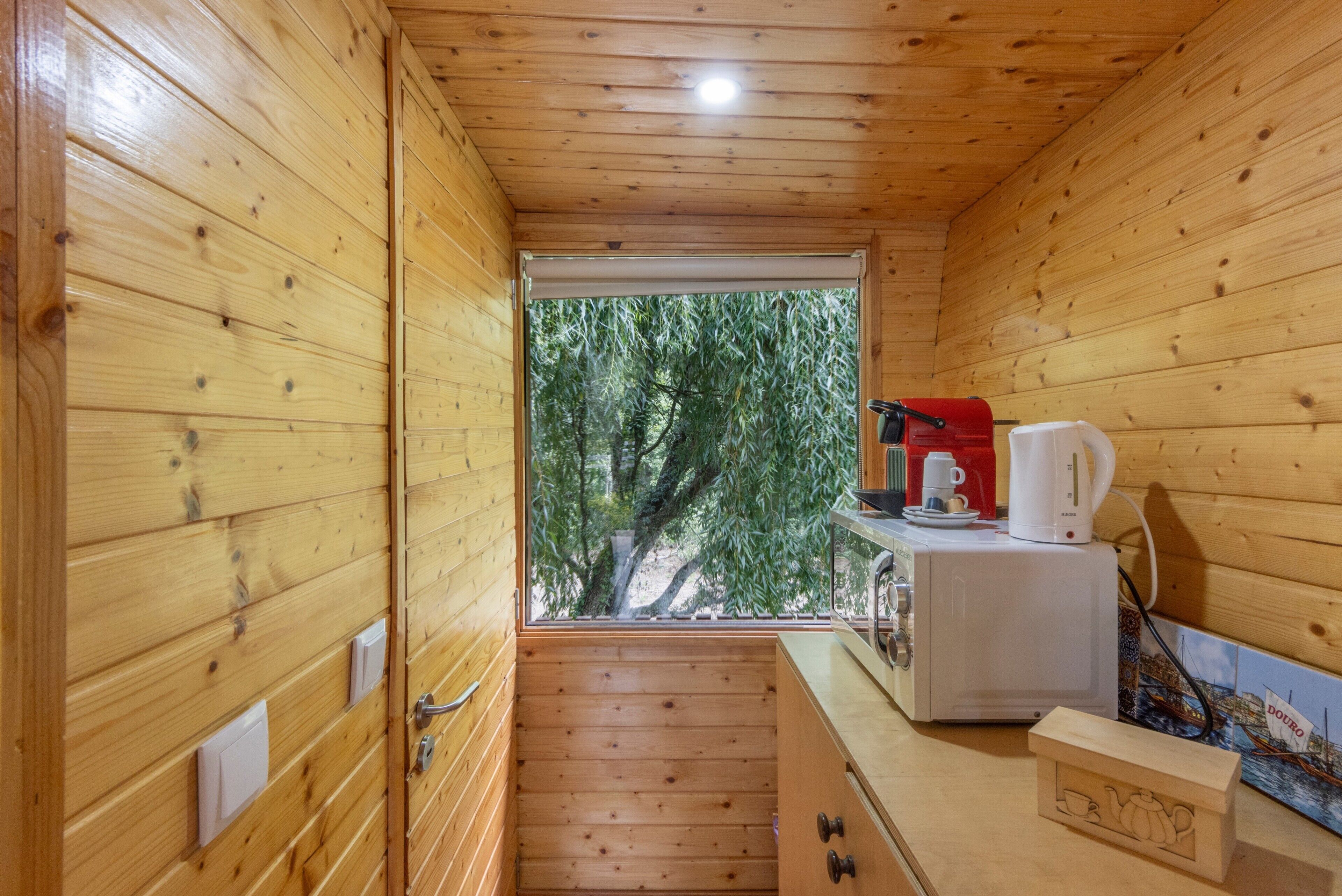 Ein gemütliches Holzhaus mit Küchenzeile, einem Fenster mit Blick auf Bäume und einem kleinen Tisch mit Wasserkocher und Tassen.