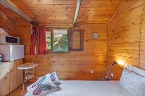 A cozy cabin room with wooden walls, a bed with patterned pillows, a small table, a microwave, and a window with a view of greenery.