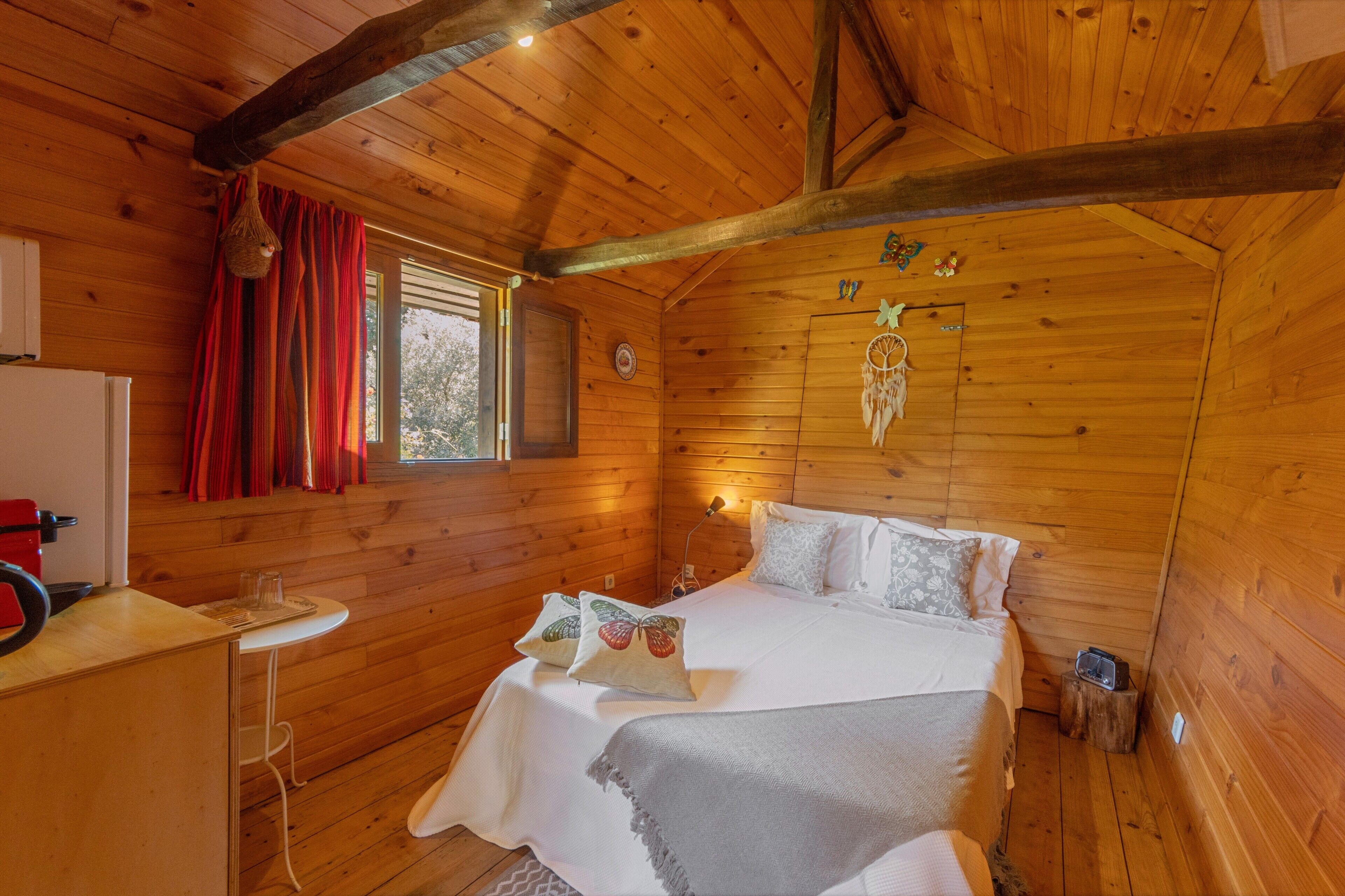 A wooden cabin bedroom with a bed, a nightstand, a lamp, and a window with curtains.