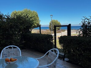 A patio with a table, chairs, and a bowl of fruit. A fence with a view of the beach and ocean is visible in the background.