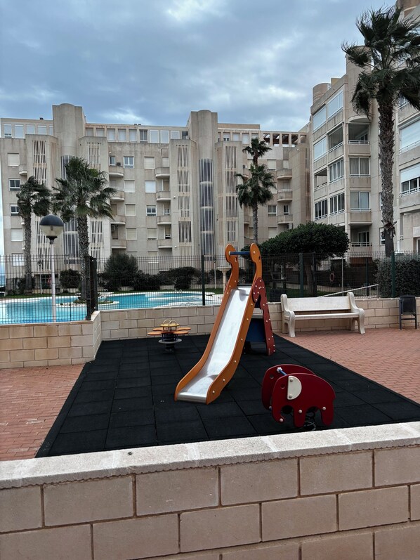 A playground with a slide and a red climbing structure, surrounded by a brick wall and palm trees, with a pool in the background.