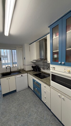 A compact kitchen with a dishwasher, stove, and a window with curtains. - Beachfront apartment with private terrace, pool and parking (El Campello)