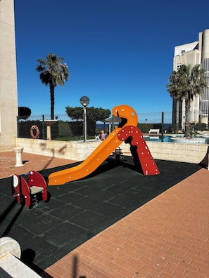 A playground with a slide and swings, surrounded by a paved area and palm trees.